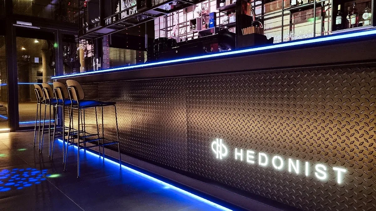 Šank s reljefnom metalnom oplatom i osvijetljenim Hedonist znakom.
